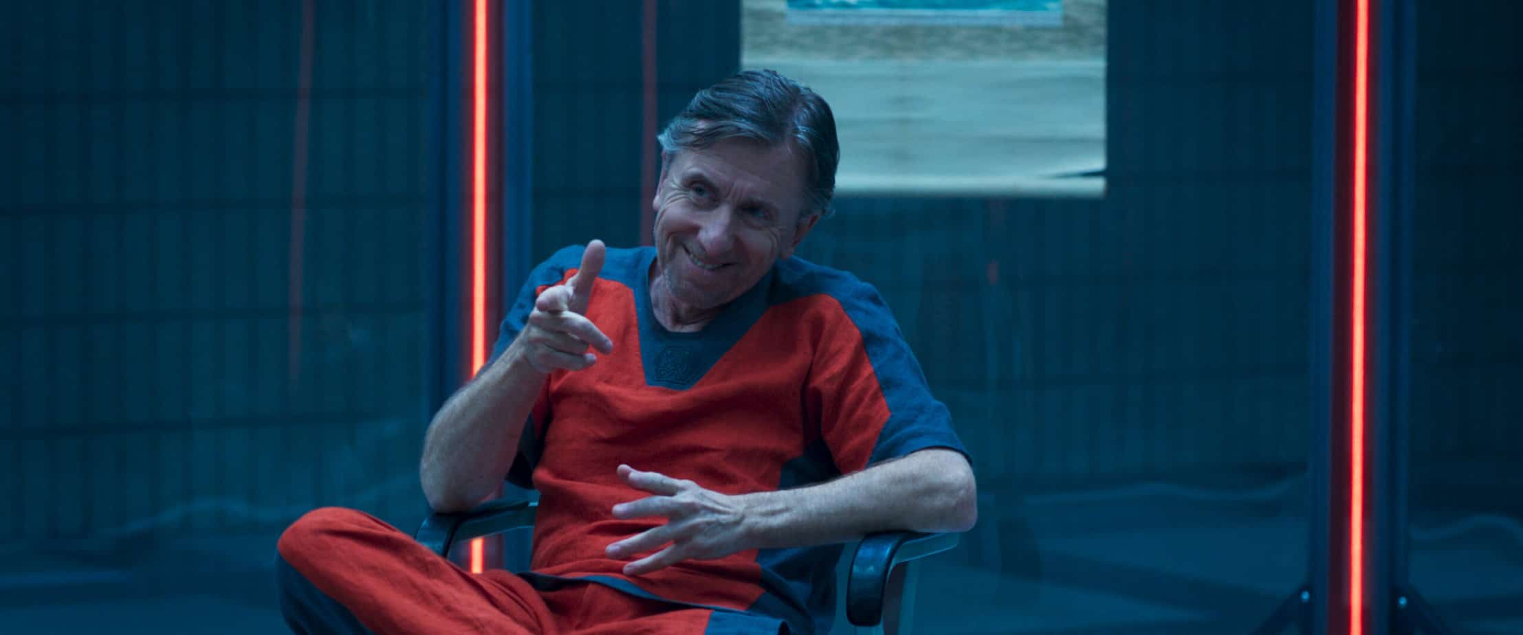 Old man in orange t-shirt and pants doing finger guns to someone off-screen. 