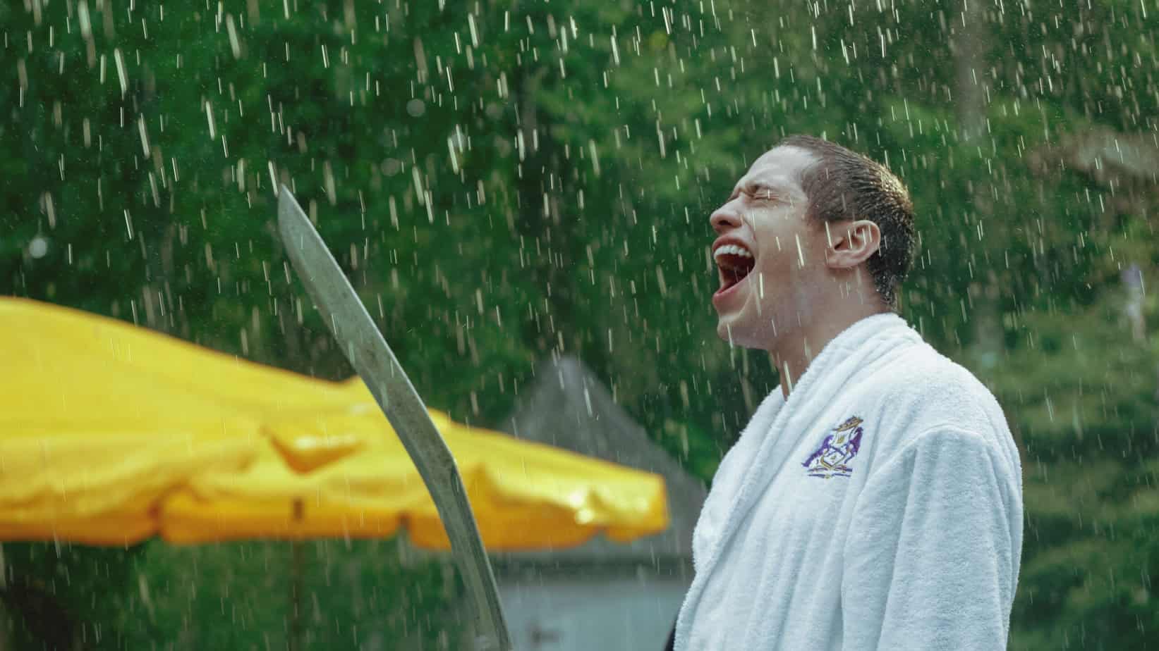 Pete DAvidson quotes from Bodies Bodies Bodies. Man standing in the rain in a robe holding a sword.