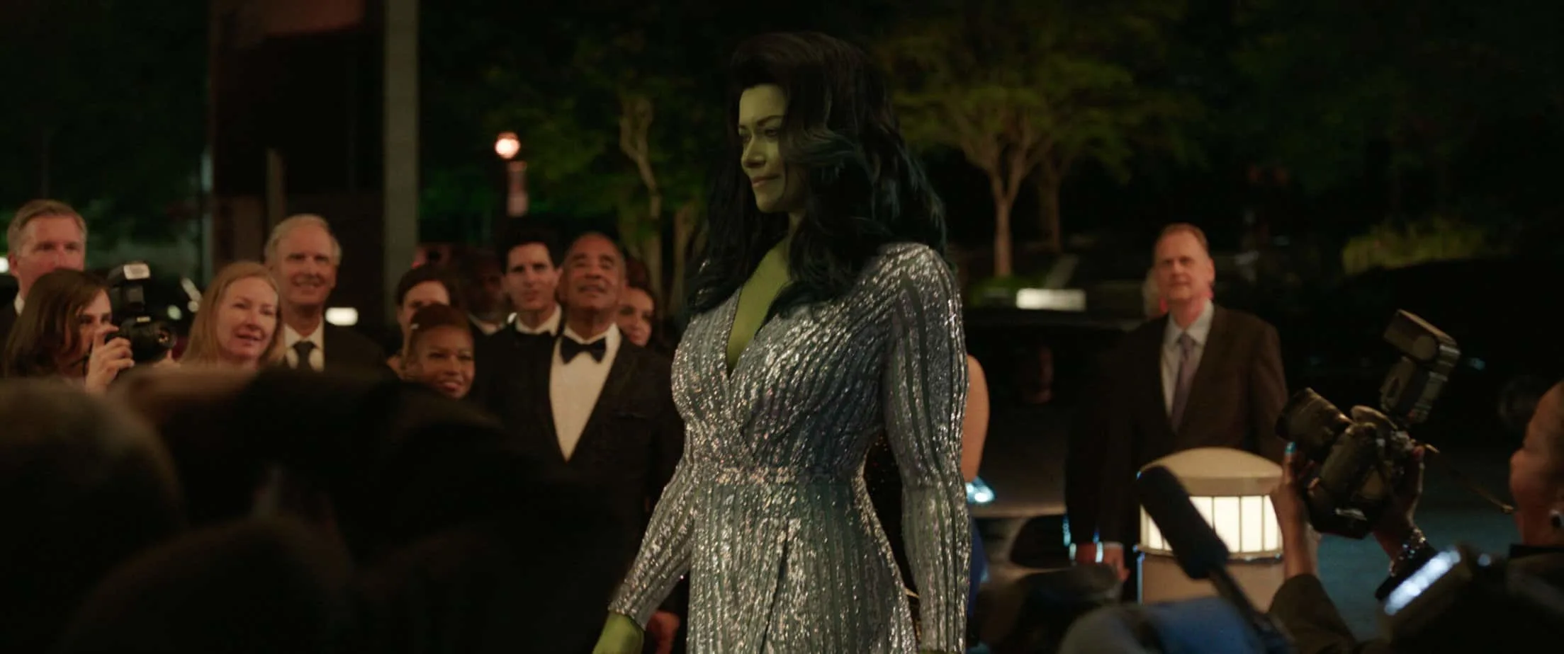 SheHulk (Big green woman) is walking through a a crowd, with everyone staring at her. She is wearing a fancy silver dress. 