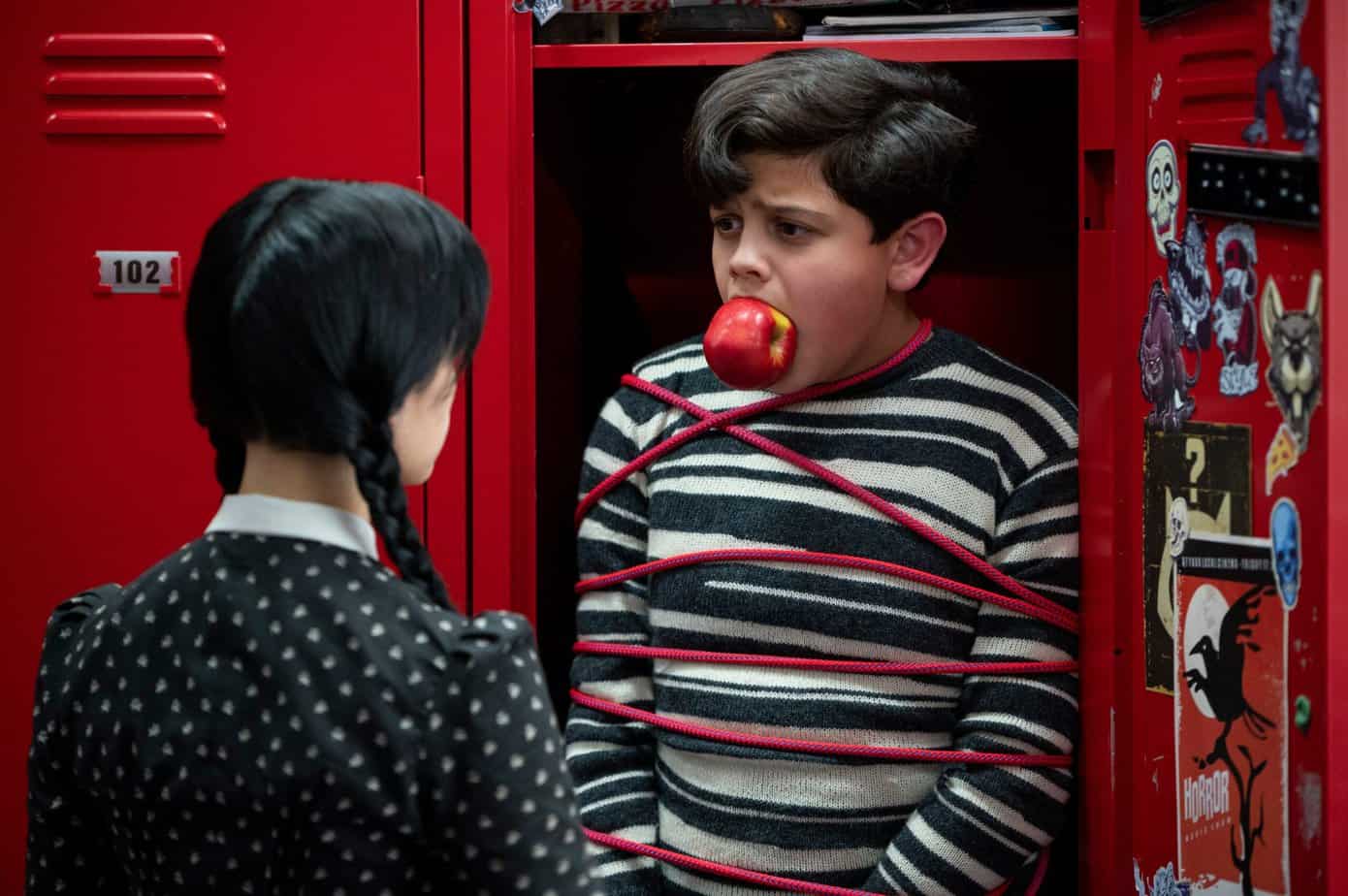 addams family quotes from Wednesday series on Netflix. Boy tied up in locker with an apple in mouth, girl looking at him.
