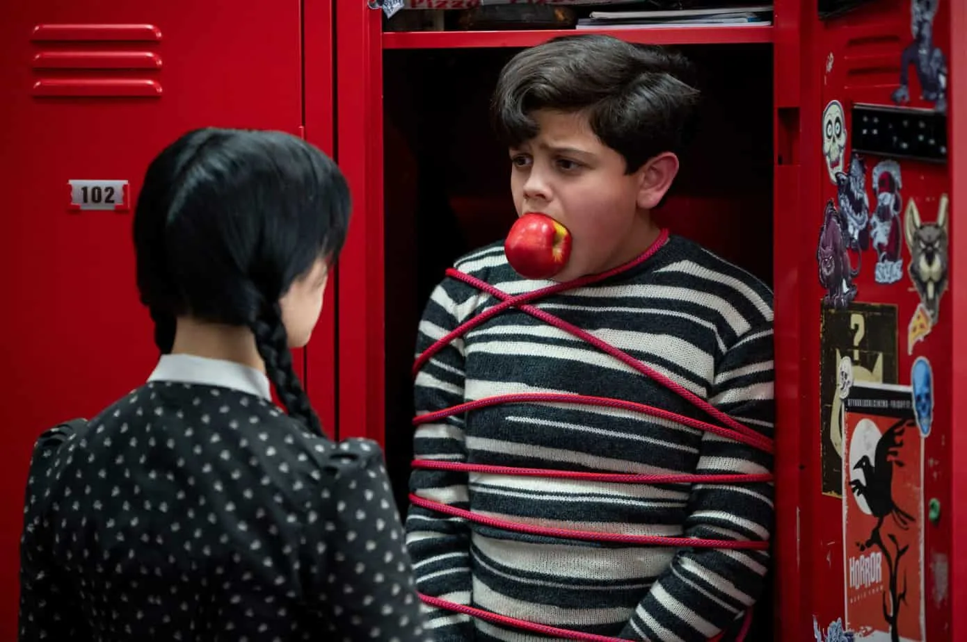 addams family quotes from Wednesday series on Netflix. Boy tied up in locker with an apple in mouth, girl looking at him.
