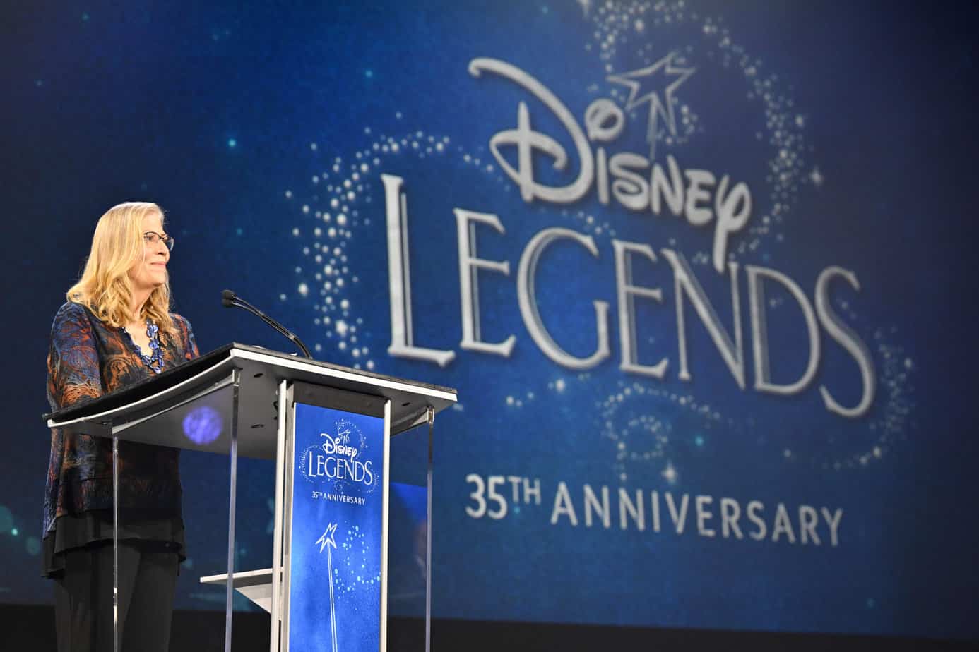 Debby Swenson speaks at a podium with the Disney Legends 35th Anniversary logo on a screen behind her. 