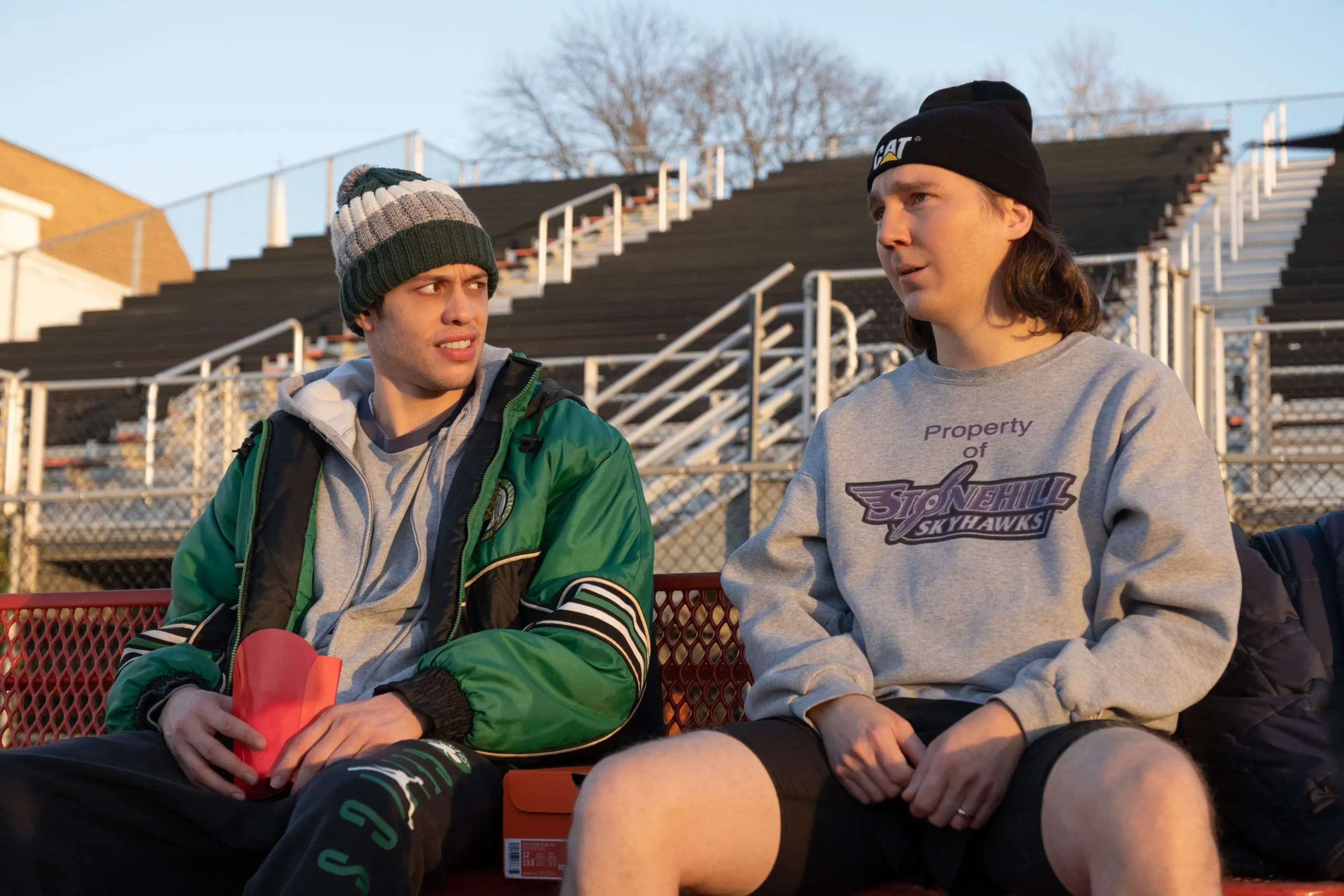 movie quotes from Dumb Money. Two guys sitting on bleachers at a high school track in the cold.